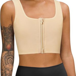 A person sporting a beige zip-front ftm chest binder and black athletic shorts stands confidently. The individual, with a tattoo sleeve on their left arm featuring intricate designs, is also wearing a discreet black binder underneath. The cropped chest binder has broad shoulder straps, set against a plain white background.