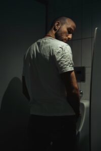 Man standing in a dark bathroom with his back facing forward, face turned back towards the camera, wearing a white shirt and dark bottom garments. The photo is dimly illuminated and the man has a suspicious, serious look on his face