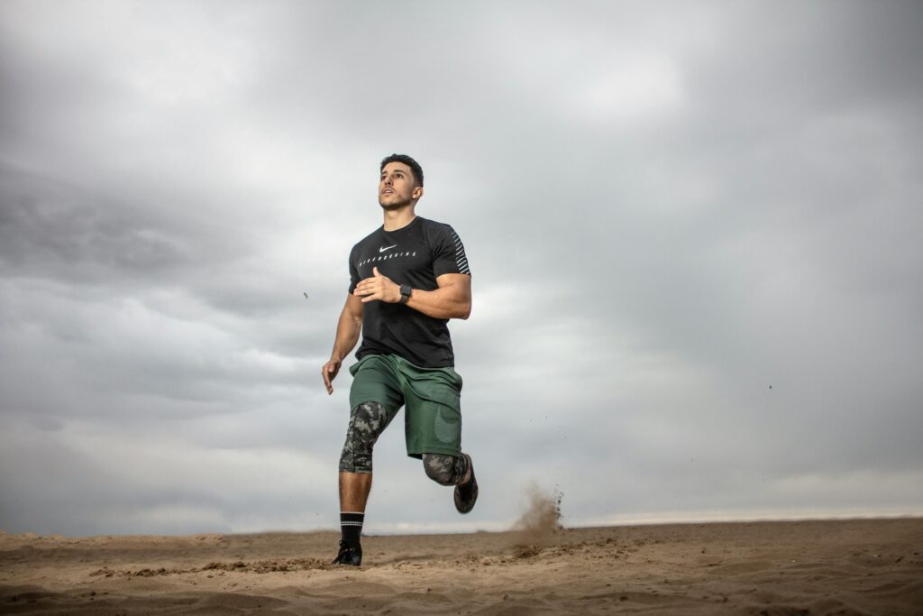 A man is running on a sandy surface under a cloudy sky. He is wearing a black t-shirt, green shorts, black and gray knee-length compression leggings, and black athletic shoes. His expression appears focused and determined as he runs.