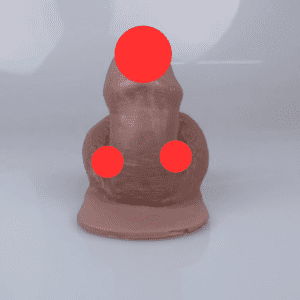 A silicone trans masculine packer is visible and sitting upright. Its smooth, brown surface showcases three red circular markers on the head, torso, and base for modesty. The backdrop remains a plain light grey.