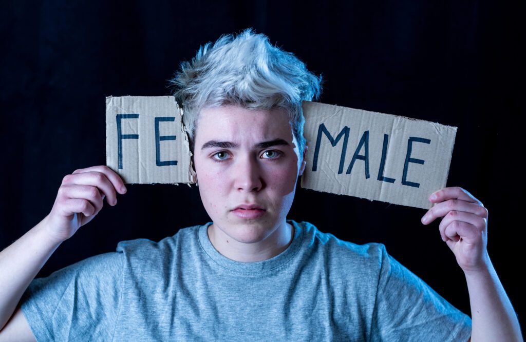 Fe male gender A person with short, light-colored hair holds two cardboard signs behind their head reading "FEMALE" and "MALE"—each by one ear—looking seriously at the camera against a dark background, highlighting questions of gender and trans identity.
