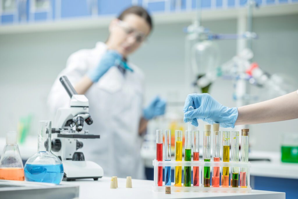 Science A lab technician wearing gloves handles test tubes with various colored liquids, while another person in a lab coat works near a microscope—highlighting the vital role of gender diversity and trans identity in advancing modern science.