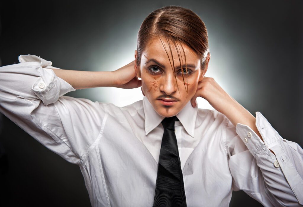 makeup man A person wearing a white dress shirt, black tie, and sporting slicked-back hair and a fake mustache poses confidently with hands behind their neck, lit dramatically from behind—a bold expression of gender and self-identity.