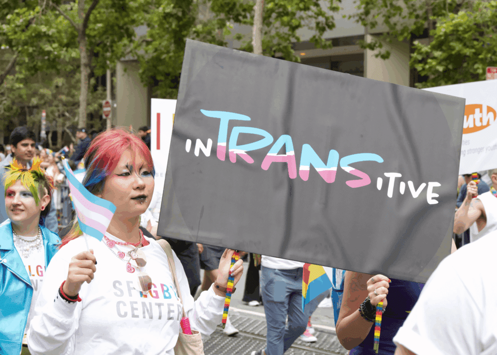 A person with colorful hair holds a transgender pride flag and a large "inTRANSitive" sign in blue, pink, and white letters at an LGBTQ pride parade, surrounded by others celebrating gay pride.