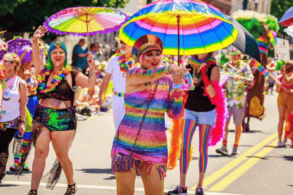 People in colorful, rainbow-themed outfits and accessories march and dance in a lively gay pride parade. Some carry rainbow umbrellas, and the street is filled with joy, celebration, and diverse LGBTQ+ opinions shared openly.