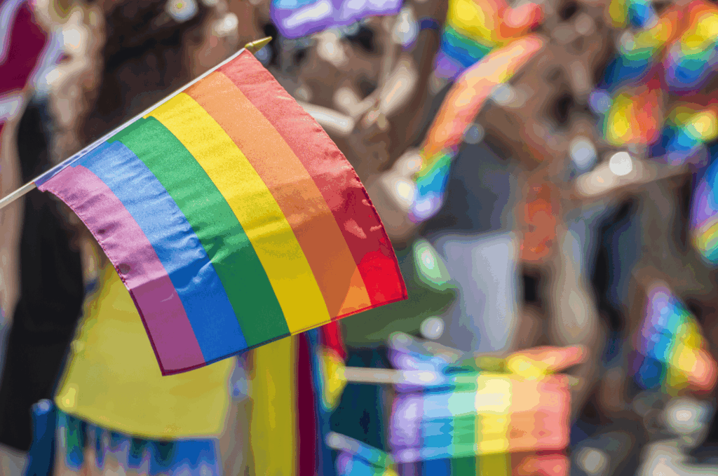 A close-up of a rainbow pride flag being waved at an outdoor event, with other rainbow flags and people in colorful clothing visible in the blurred background, captures the vibrant spirit of pride parades and celebrates gay pride.