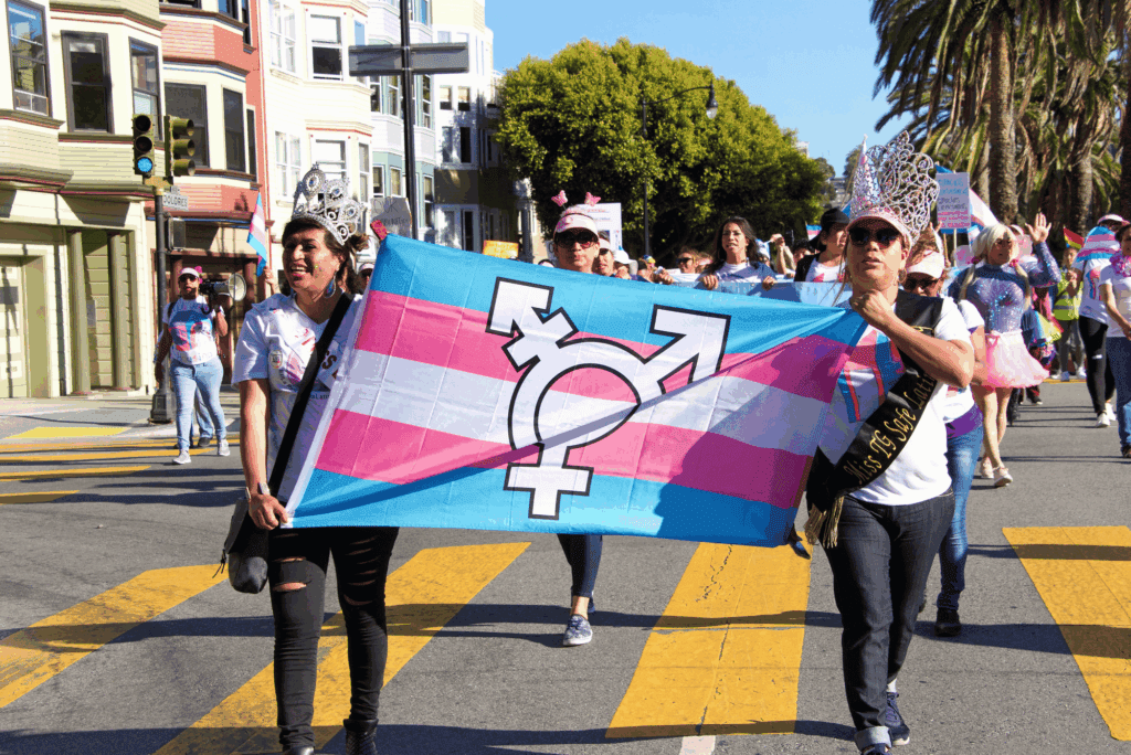 People march in a pride parade, holding a large transgender pride flag with an inclusive gender symbol. Some LGBT participants wear crowns and sashes as the group walks down a city street lined with palm trees and colorful buildings.