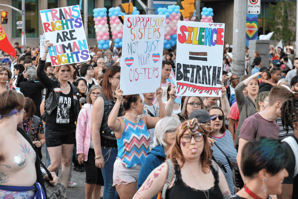 A large crowd marches at an LGBTQ+ pride event. Three people hold signs reading "TRANS RIGHTS ARE HUMAN RIGHTS," "SUPPORT YOUR SISTERS NOT JUST YOUR CIS-TERS," and "SILENCE = BETRAYAL." Balloons and rainbow flags are visible.