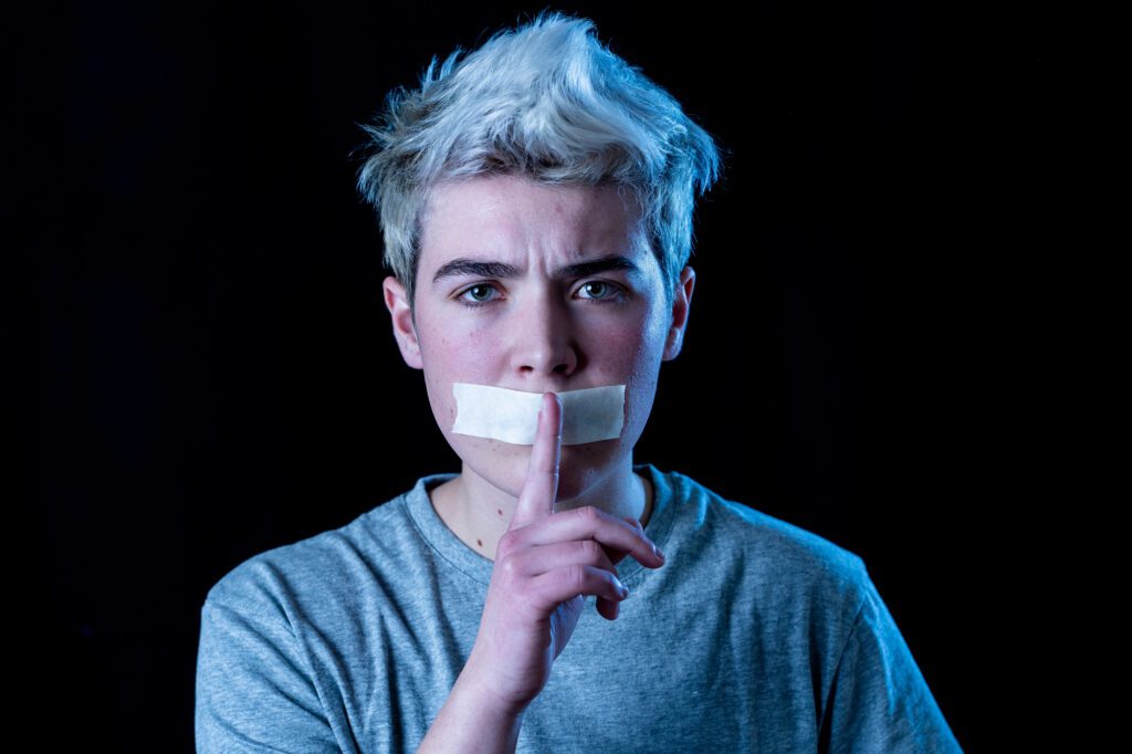 Masculine presenting person with short, light hair and gray shirt has tape over their mouth and holds a finger to their lips, respectfully signaling silence. The background is black, and the lighting casts a blue tint on one side of their face, inviting questions about identity or expression—such as those experienced by trans men.