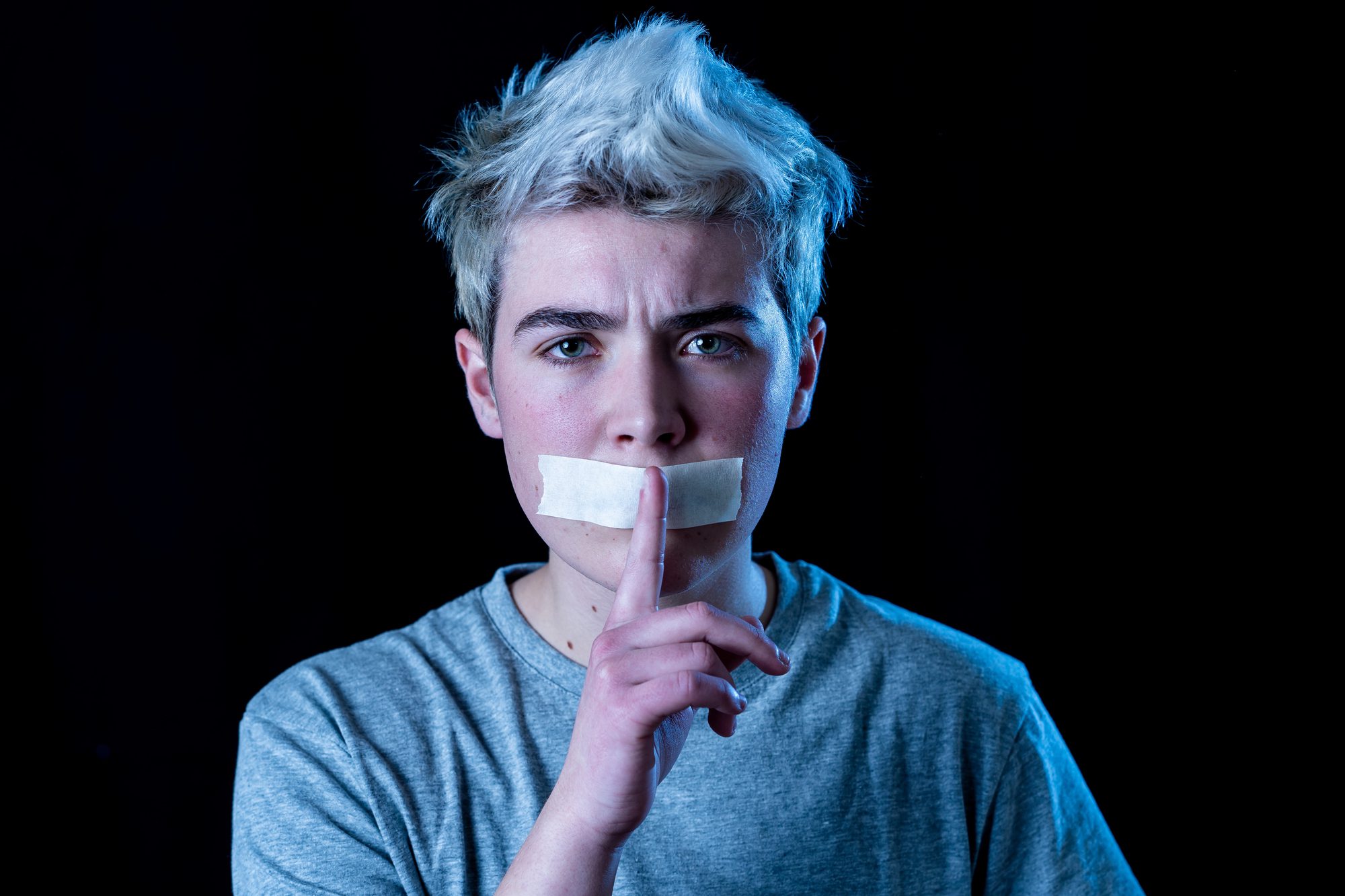Masculine presenting person with short, light hair and gray shirt has tape over their mouth and holds a finger to their lips, respectfully signaling silence. The background is black, and the lighting casts a blue tint on one side of their face, inviting questions about identity or expression—such as those experienced by trans men.