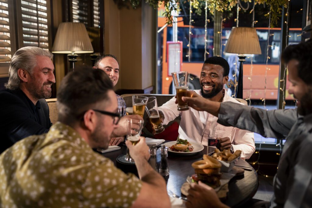 Male friends Five men sit around a restaurant table, smiling and raising glasses for a toast. Plates of food, including burgers, are on the table. Warm lighting and a window showing a bus outside create a cozy, cheerful atmosphere—perfect for sharing stories and deepening understanding by asking thoughtful questions to ask one another about their experiences as trans men.