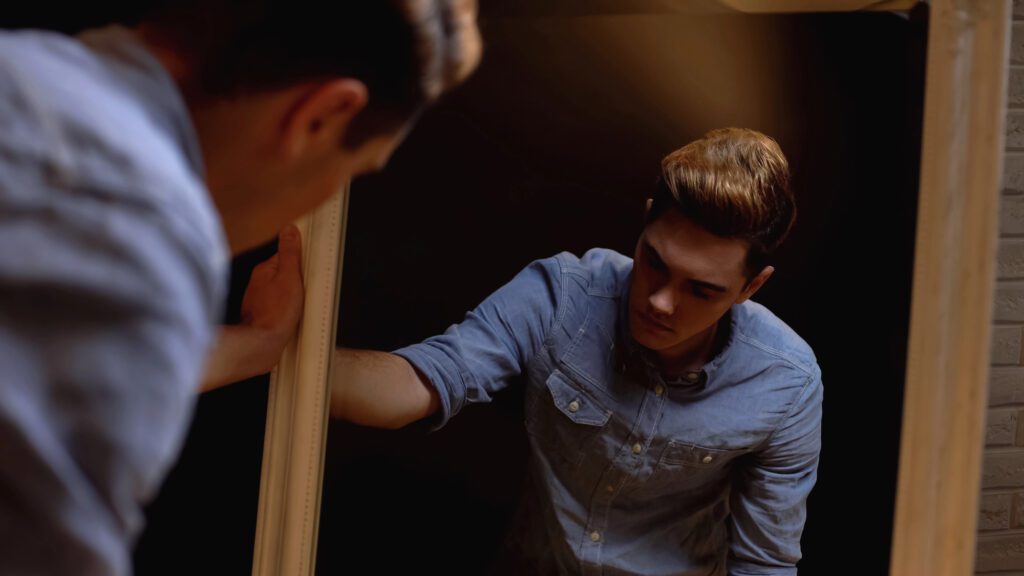 Trans in mirror A masculine person in a blue shirt leans on a mirror, looking down with a serious expression, as if lost in questions. His reflection is visible, and the lighting is dim, respectfully capturing a moment of introspection often experienced by trans men.