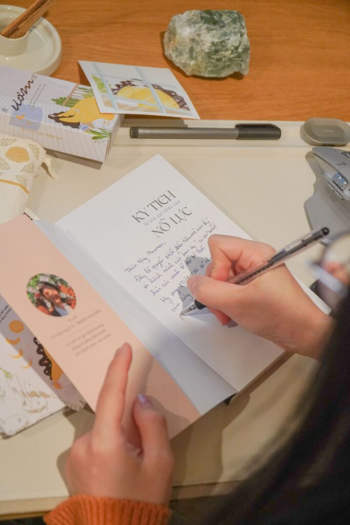 Person writing in an open journal at a wooden desk surrounded by stationery, books, and art supplies, representing the process of finding language for one’s gender identity.