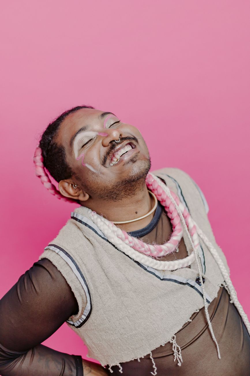 Smiling man with short dark hair and facial hair leaning backward with his eyes closed against a bright pink background, wearing layered necklaces and a sleeveless beige top.