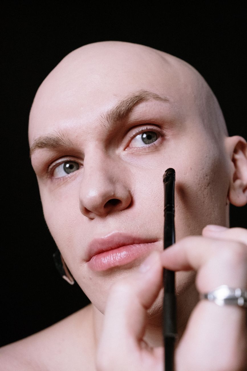 Close-up portrait of a bald person applying makeup near their eye with a brush against a black background.