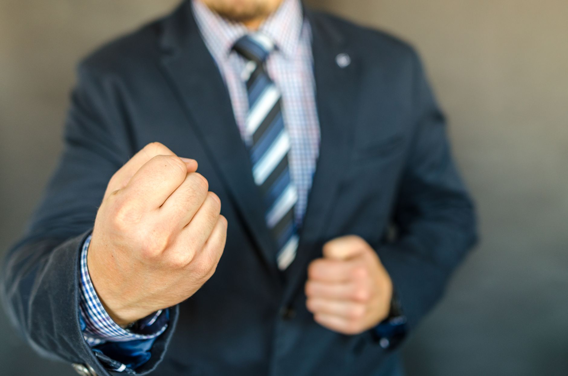 Close-up of a man wearing a dark blazer, patterned shirt, and striped tie, with his hands raised near the front of his jacket.