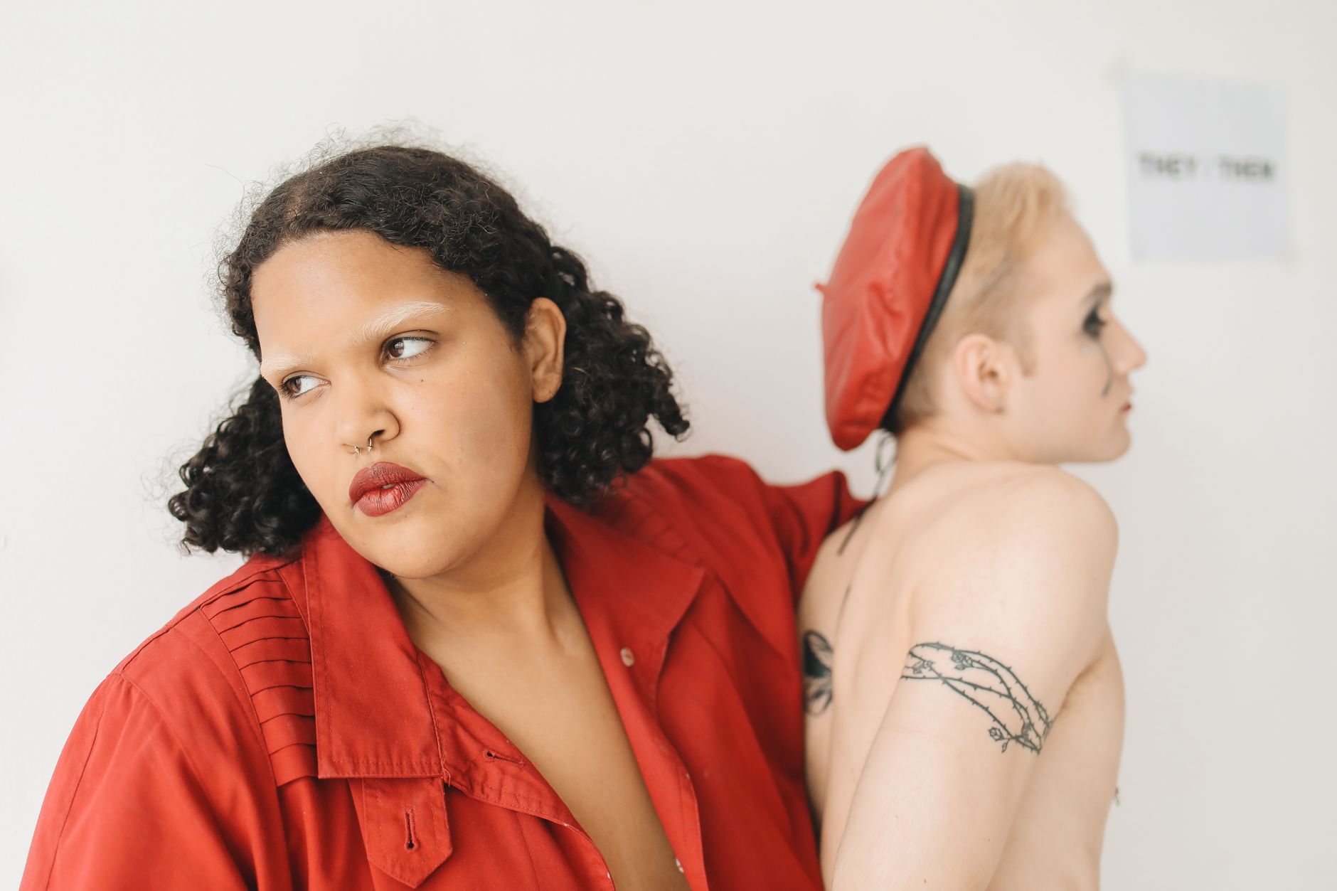 Two people posing back-to-back against a white background, one wearing a red shirt and the other shirtless with a red beret and an upper-arm tattoo.