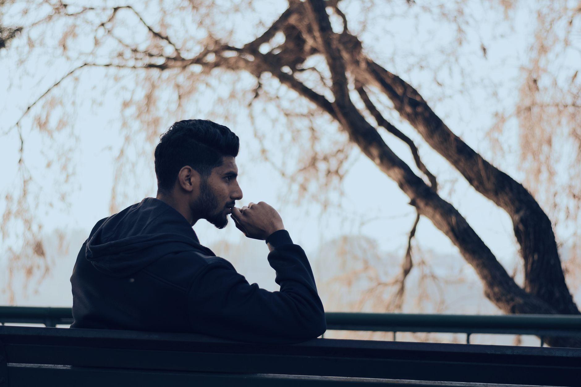 Man in a black hoodie sitting alone on a park bench beneath a tree, resting his chin on his hand while looking thoughtfully into the distance.