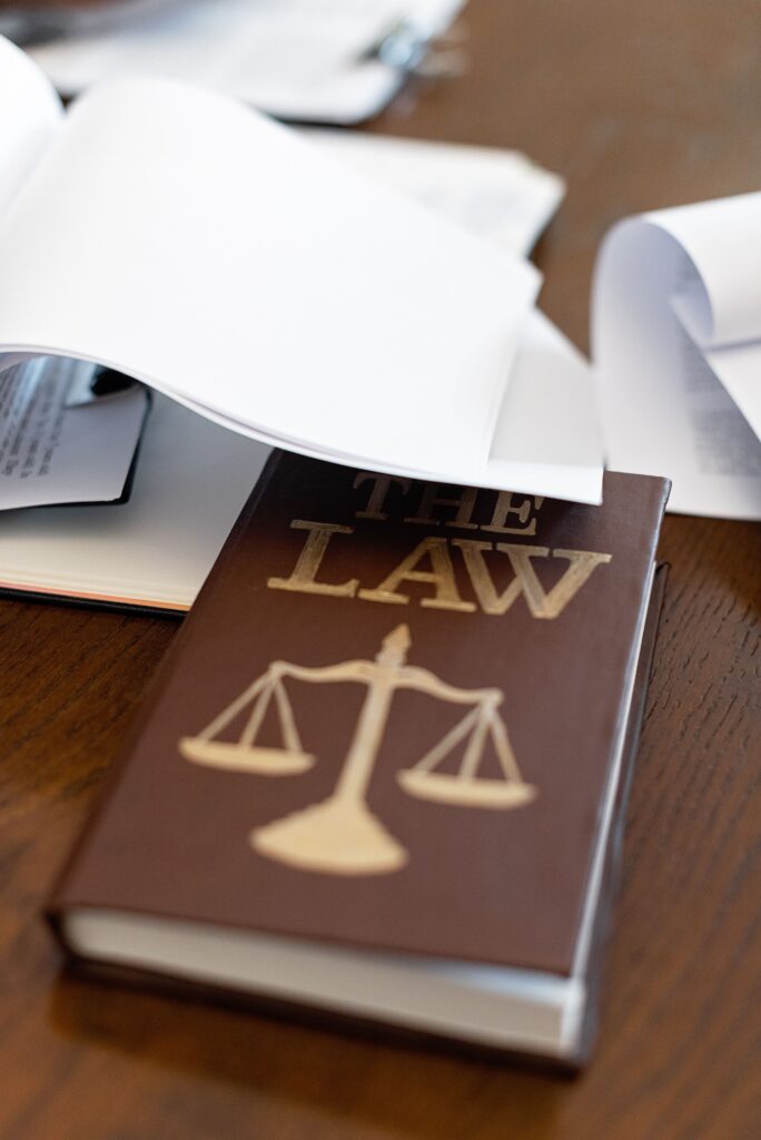 pexels-photo-8731036-8731036 Close-up of a law book with scales icon on a wooden desk covered in papers.