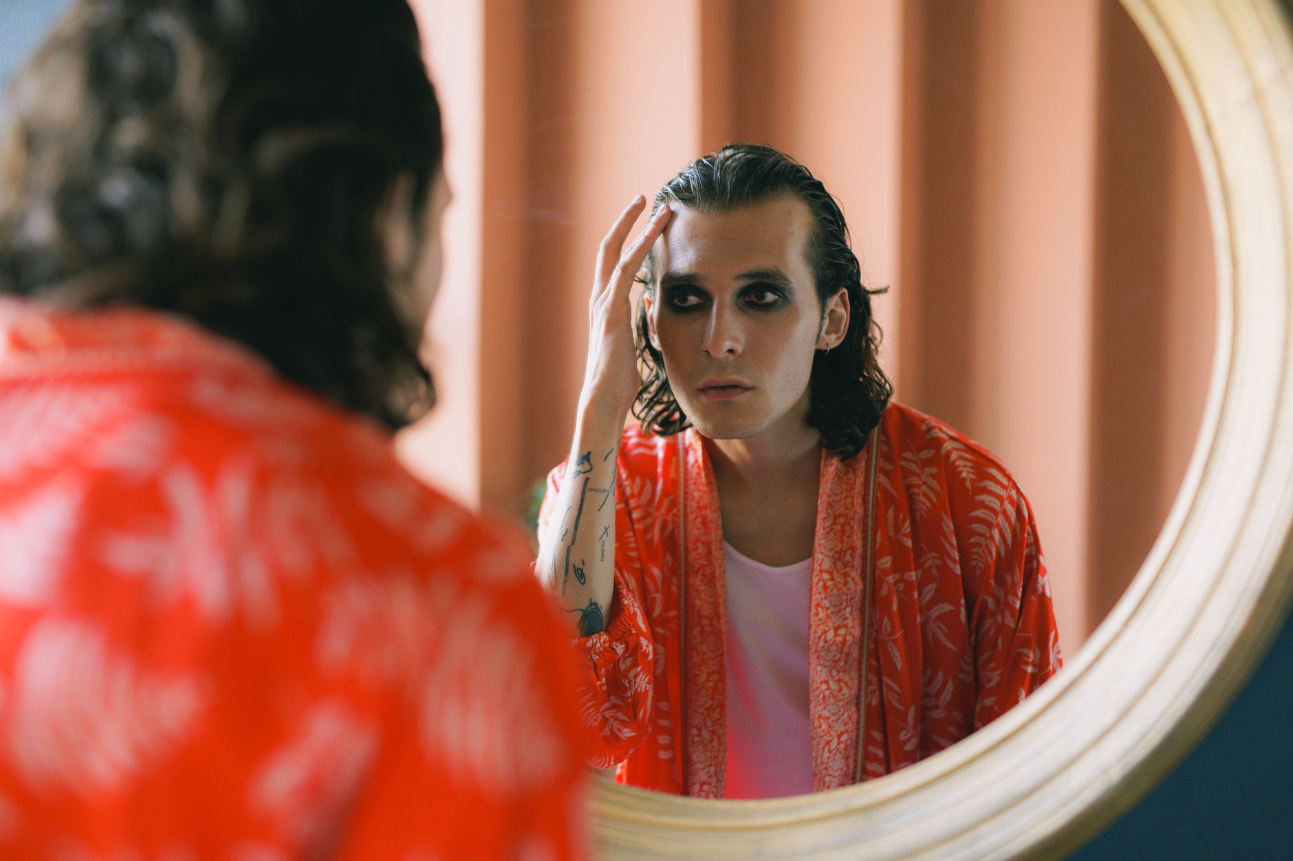 Person with dark hair and eye makeup looking at their reflection in a round mirror while wearing a red patterned garment.