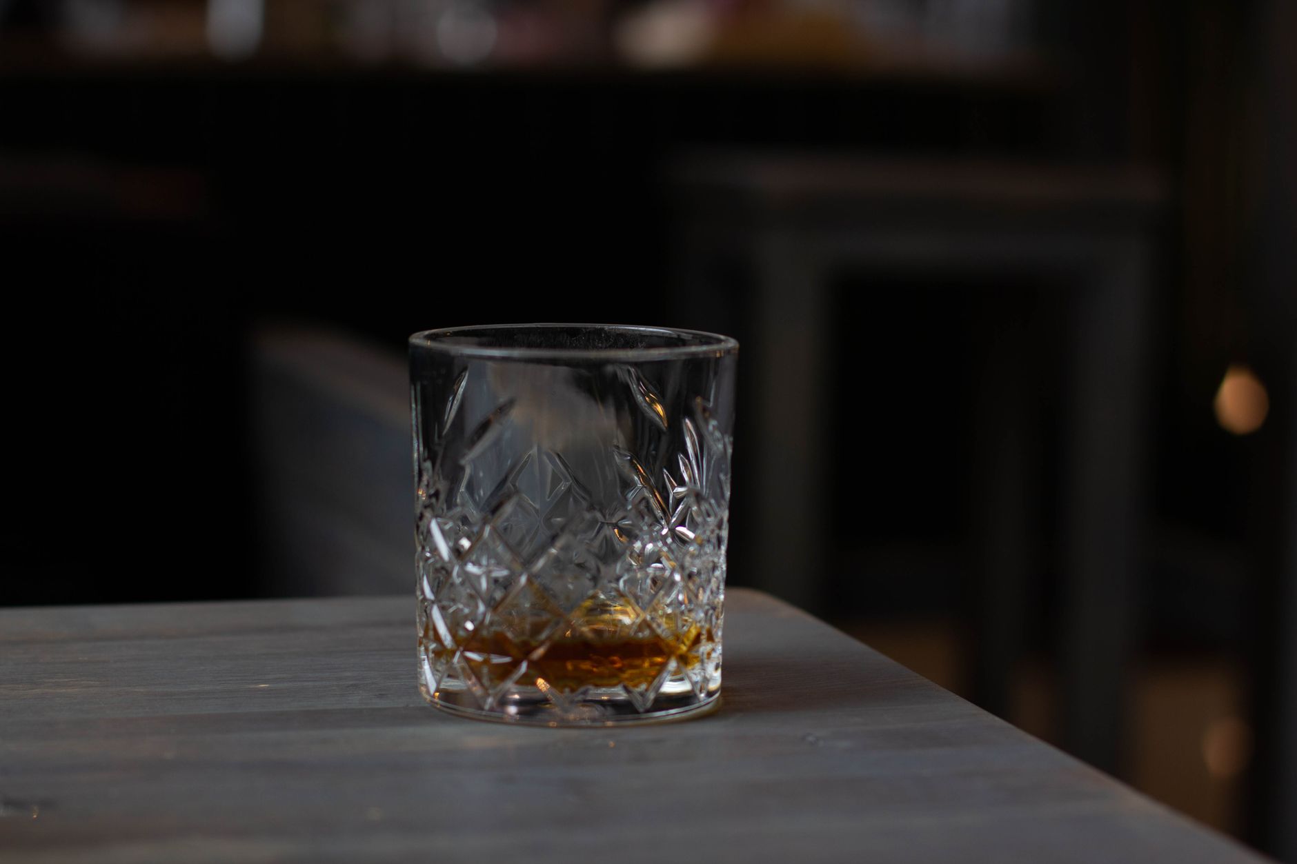 A nearly empty whiskey glass with a small amount of amber liquor sits on a wooden table in a dimly lit room.